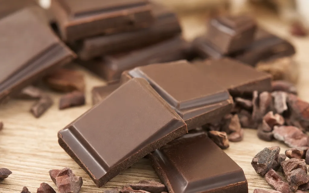 Carrés de chocolat noir posés sur une table en bois clair, lumière naturelle douce, fond crème épuré